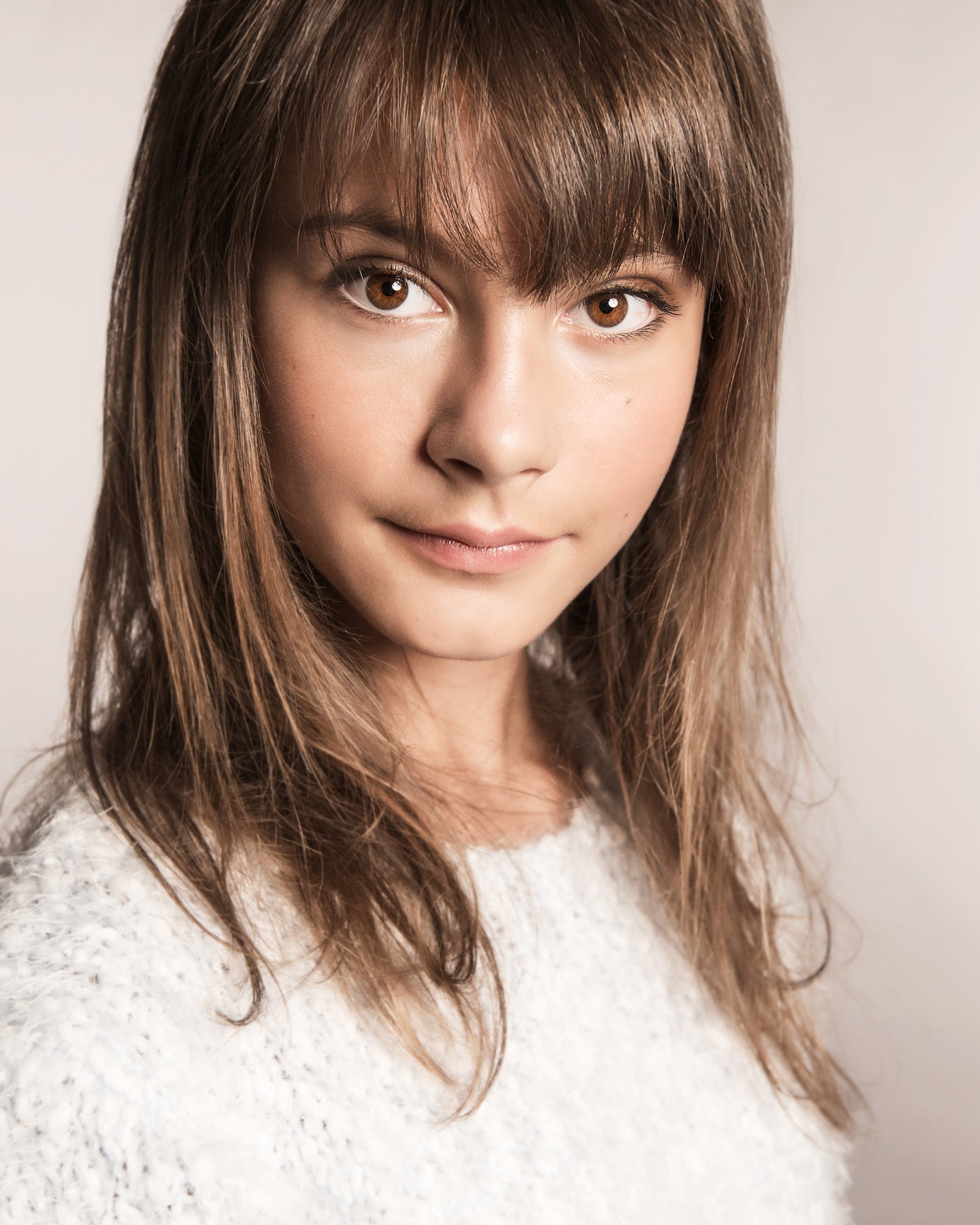 Young woman with long brown hair and bangs wearing a white sweater against a plain background
