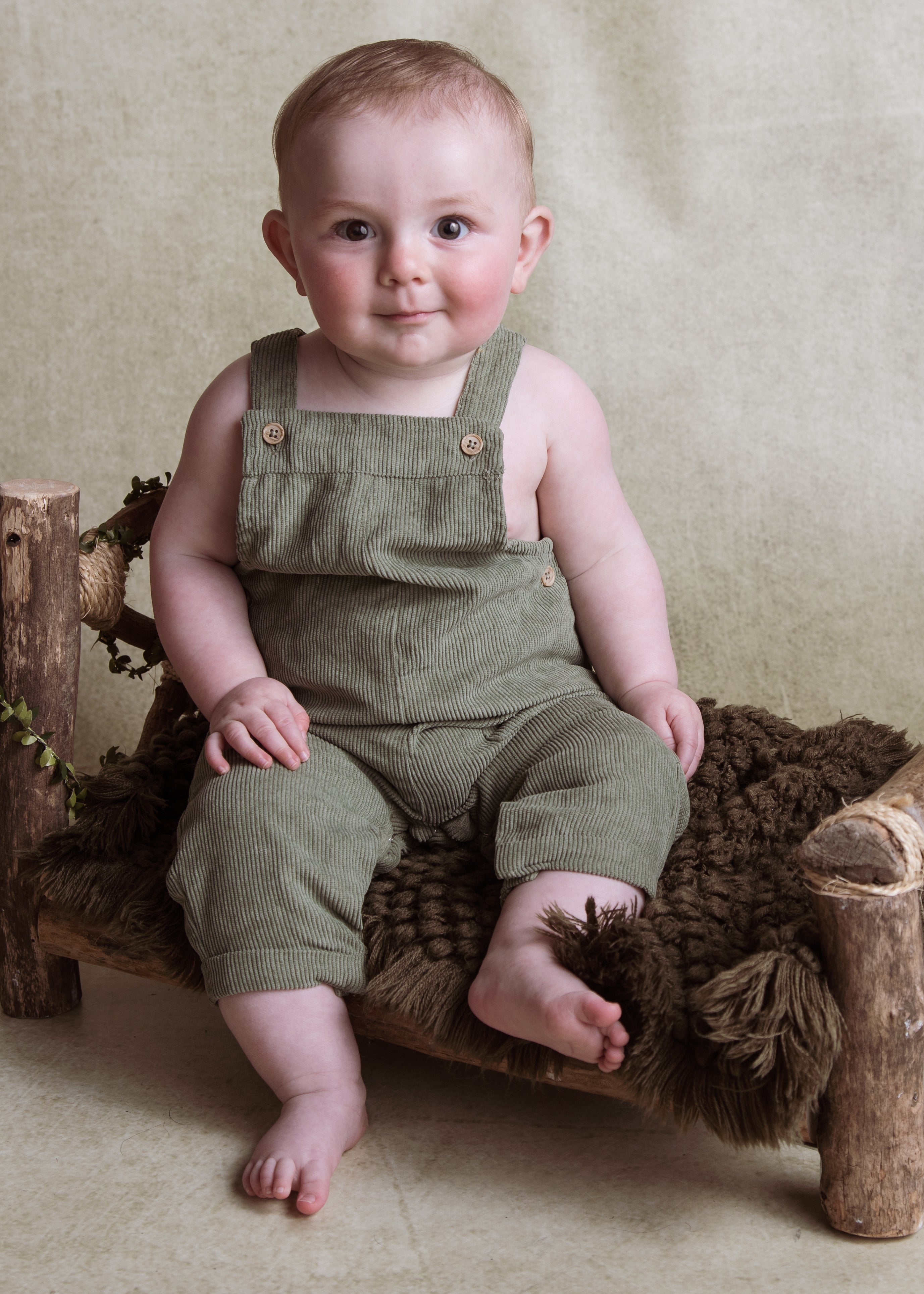 Baby in green overalls sitting on a wooden bench with a textured beige background