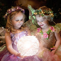Two children in fairy costumes holding a glowing magical orb in a woodland setting.