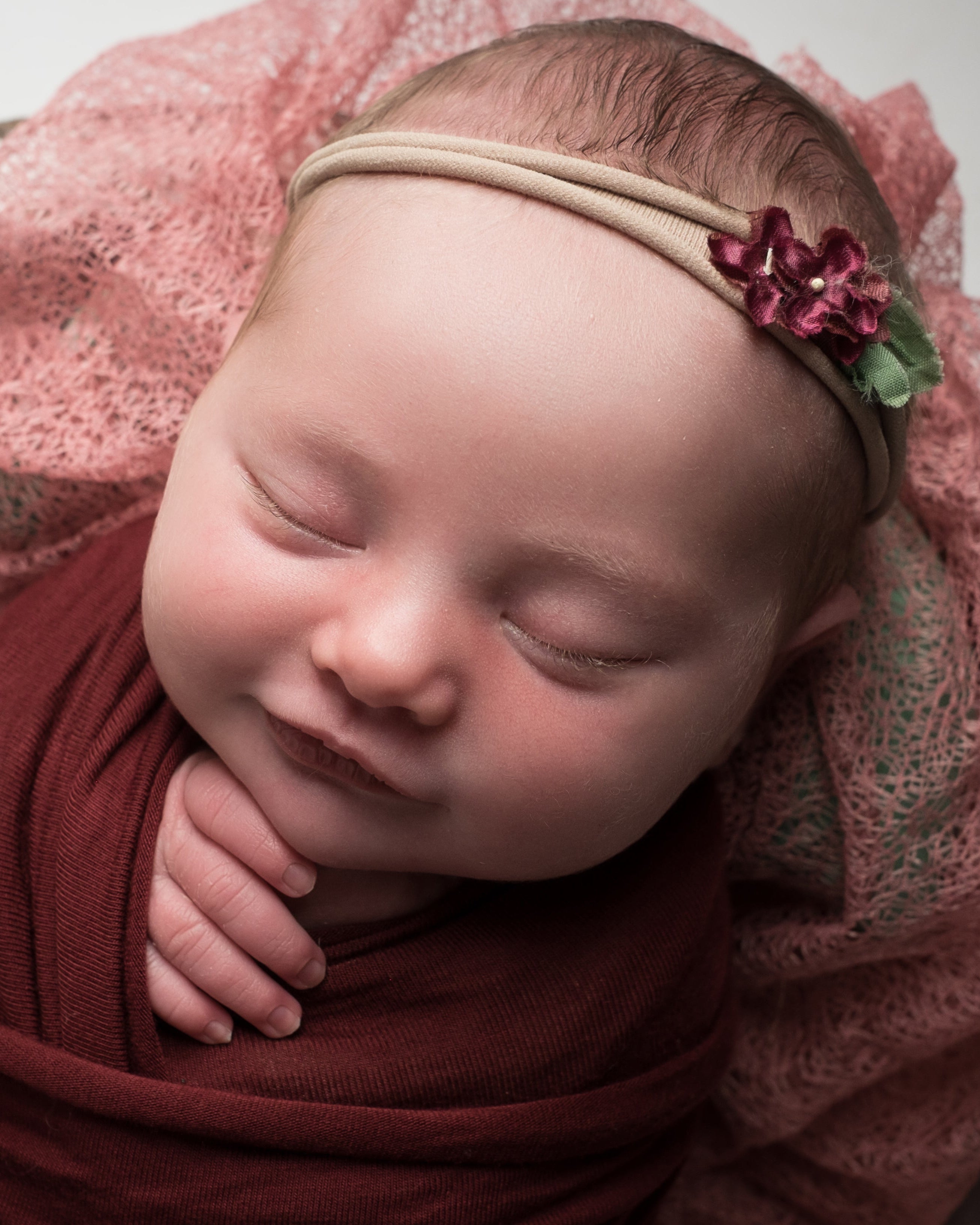 Newborn baby wrapped in red fabric with a pink headband, lying on a textured surface.