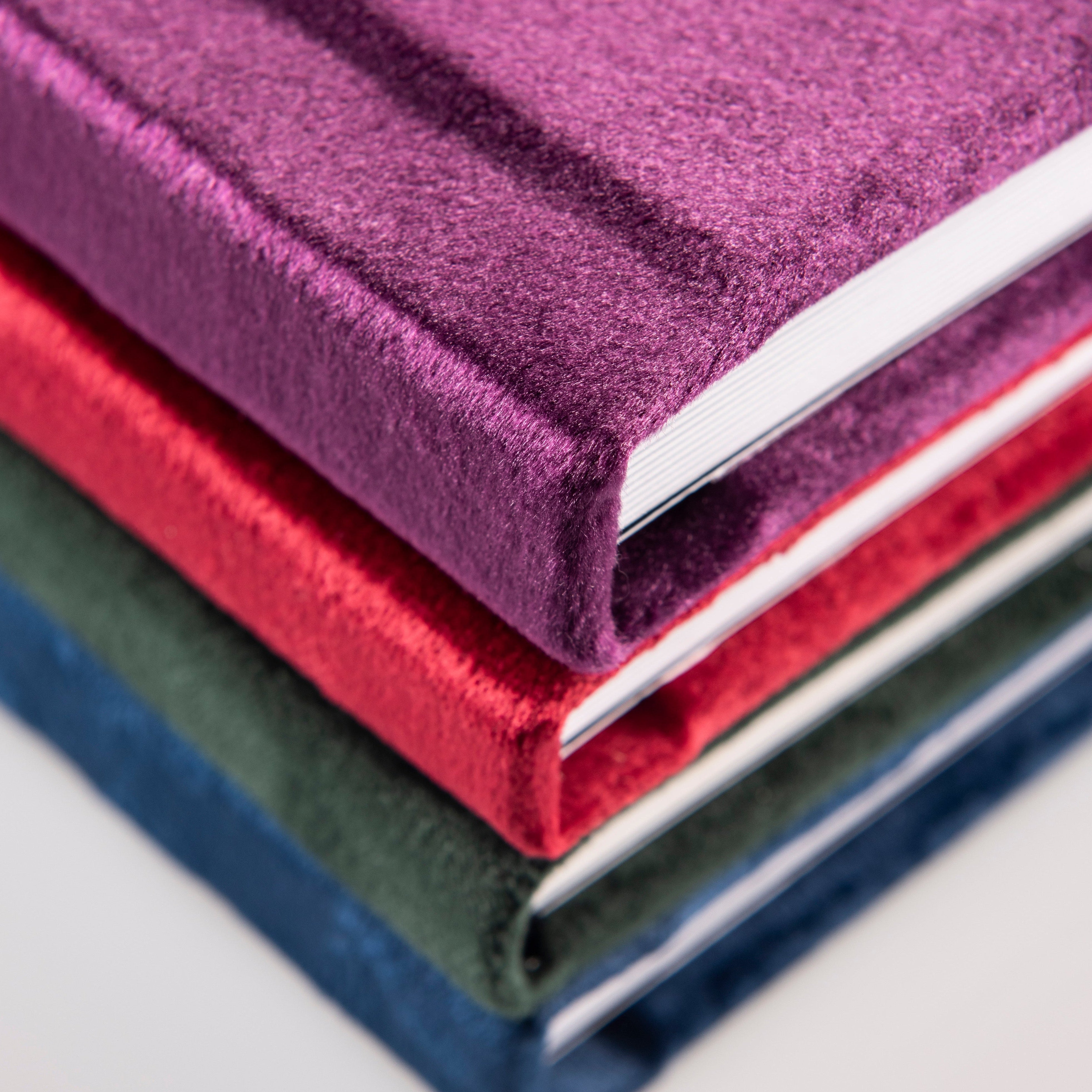 Stack of three fabric-bound books in purple, red, and green on a light gray background