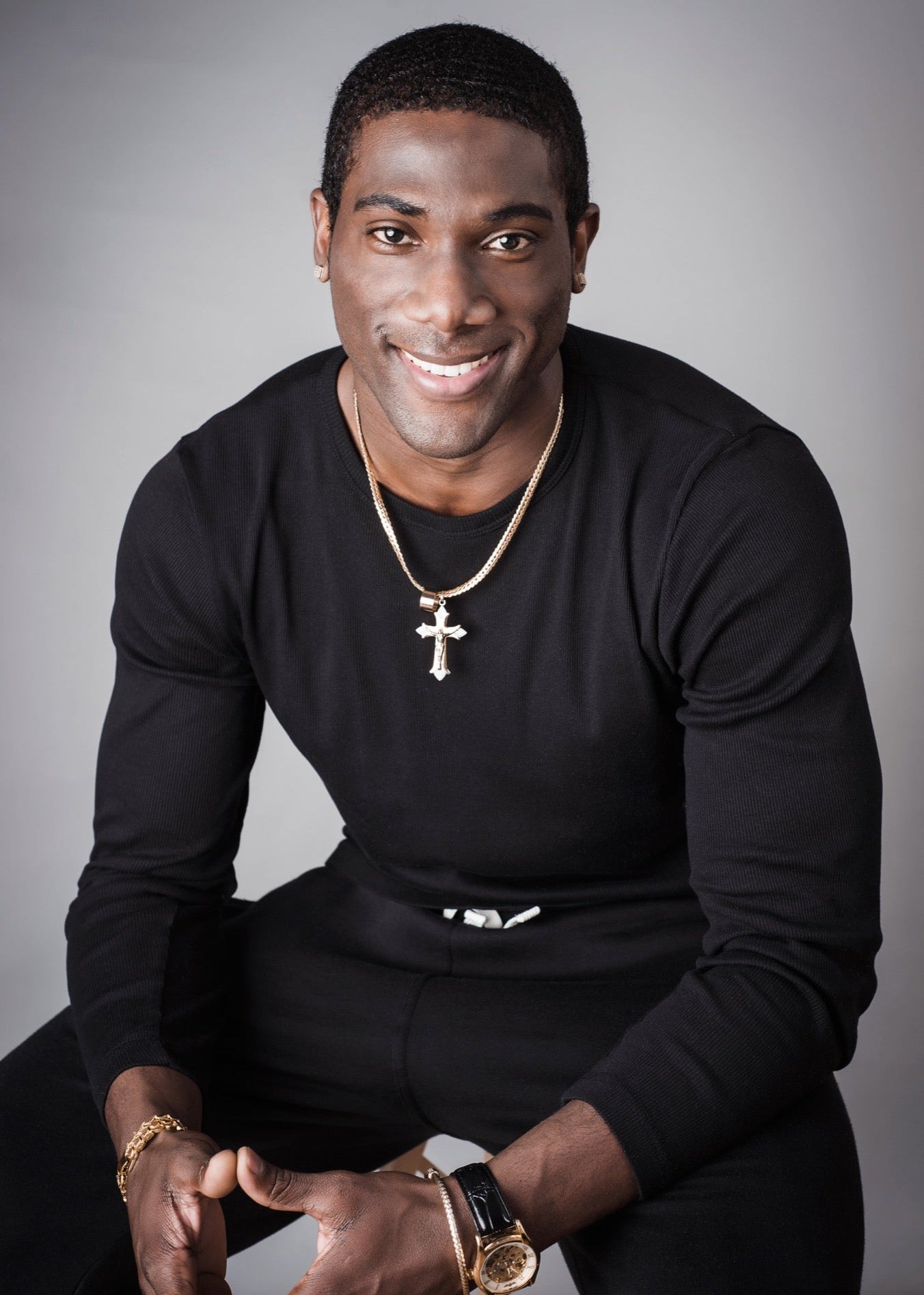 Man wearing a black long-sleeve shirt with a gold necklace and watch, sitting against a gray background.